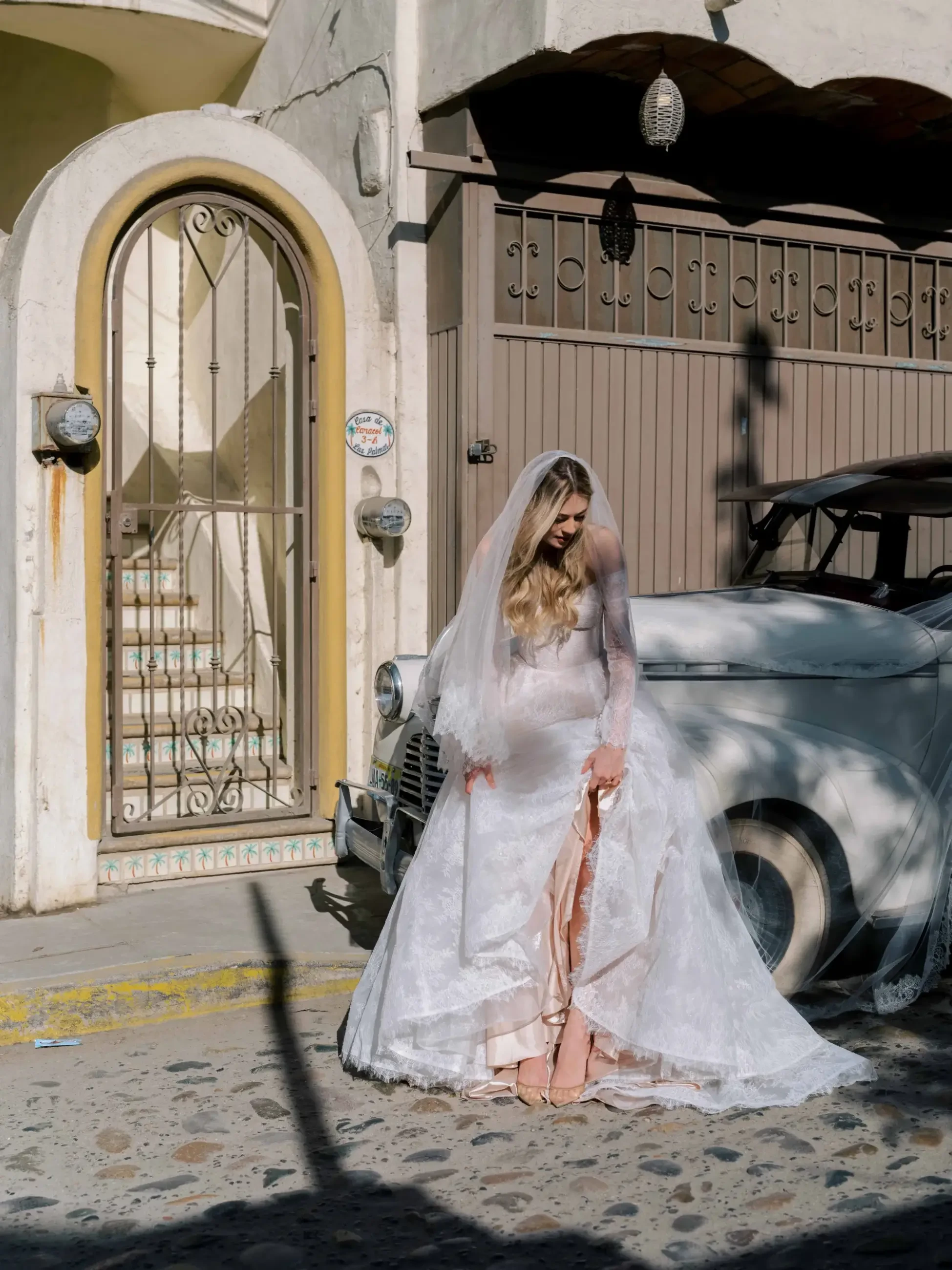 A bride in a white lace gown and veil stands beside a vintage car on a cobblestone street, looking down. Sunlight casts soft shadows, creating a serene mood.
