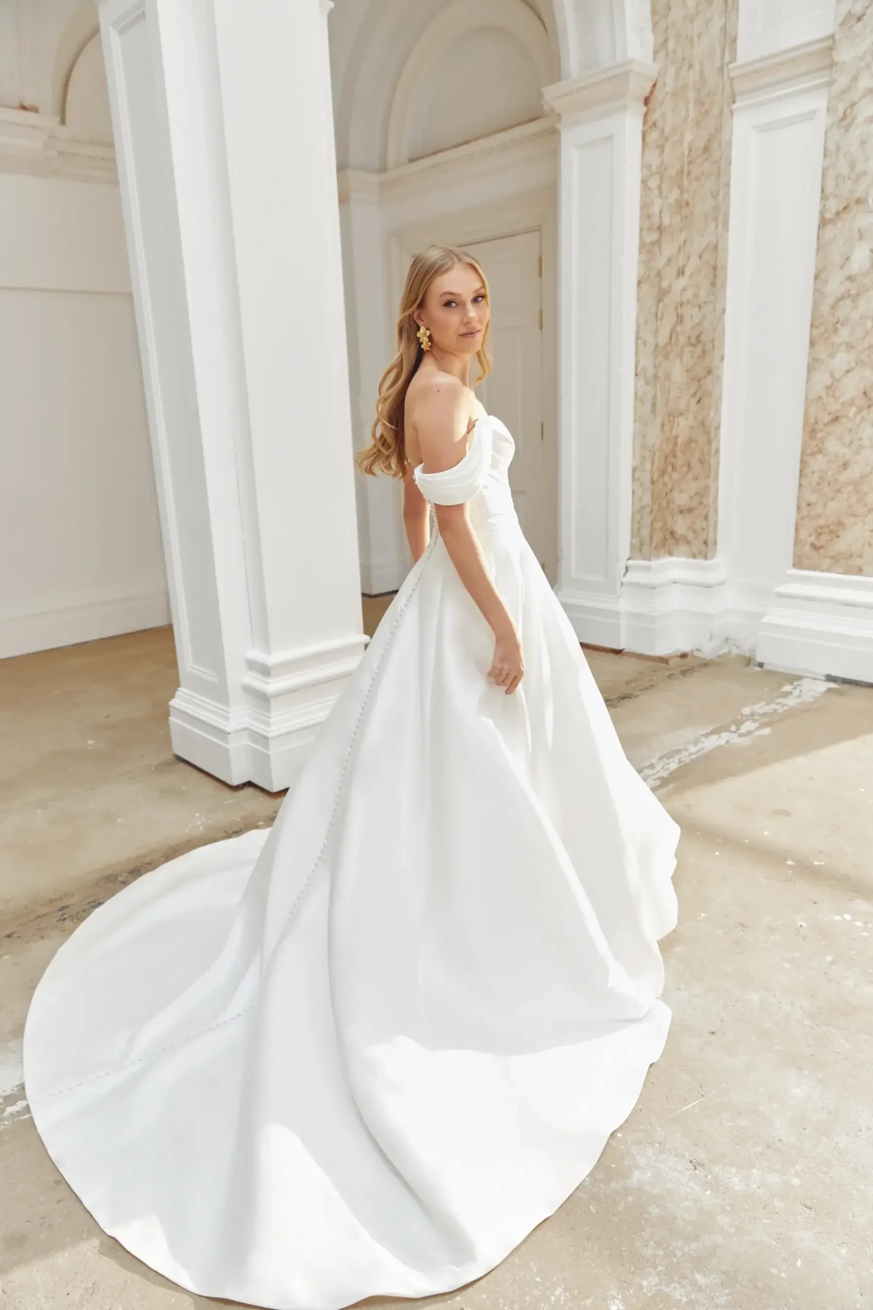 A woman in an elegant off-the-shoulder white wedding gown stands in a grand hall with high ceilings and classical columns, exuding grace and poise.