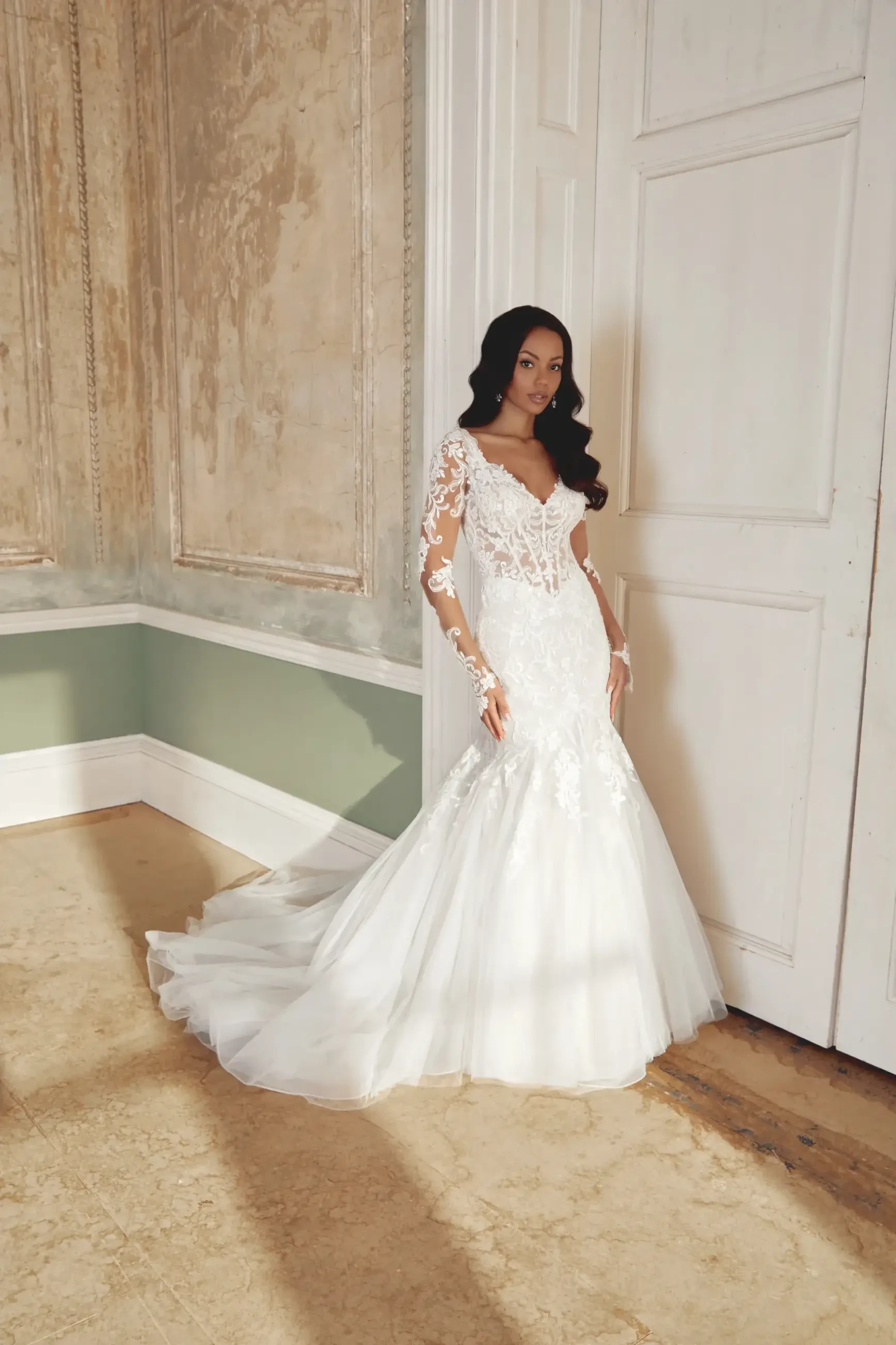 A woman in an elegant white wedding dress with lace details, standing in a softly lit room with wooden walls and a light-colored floor.