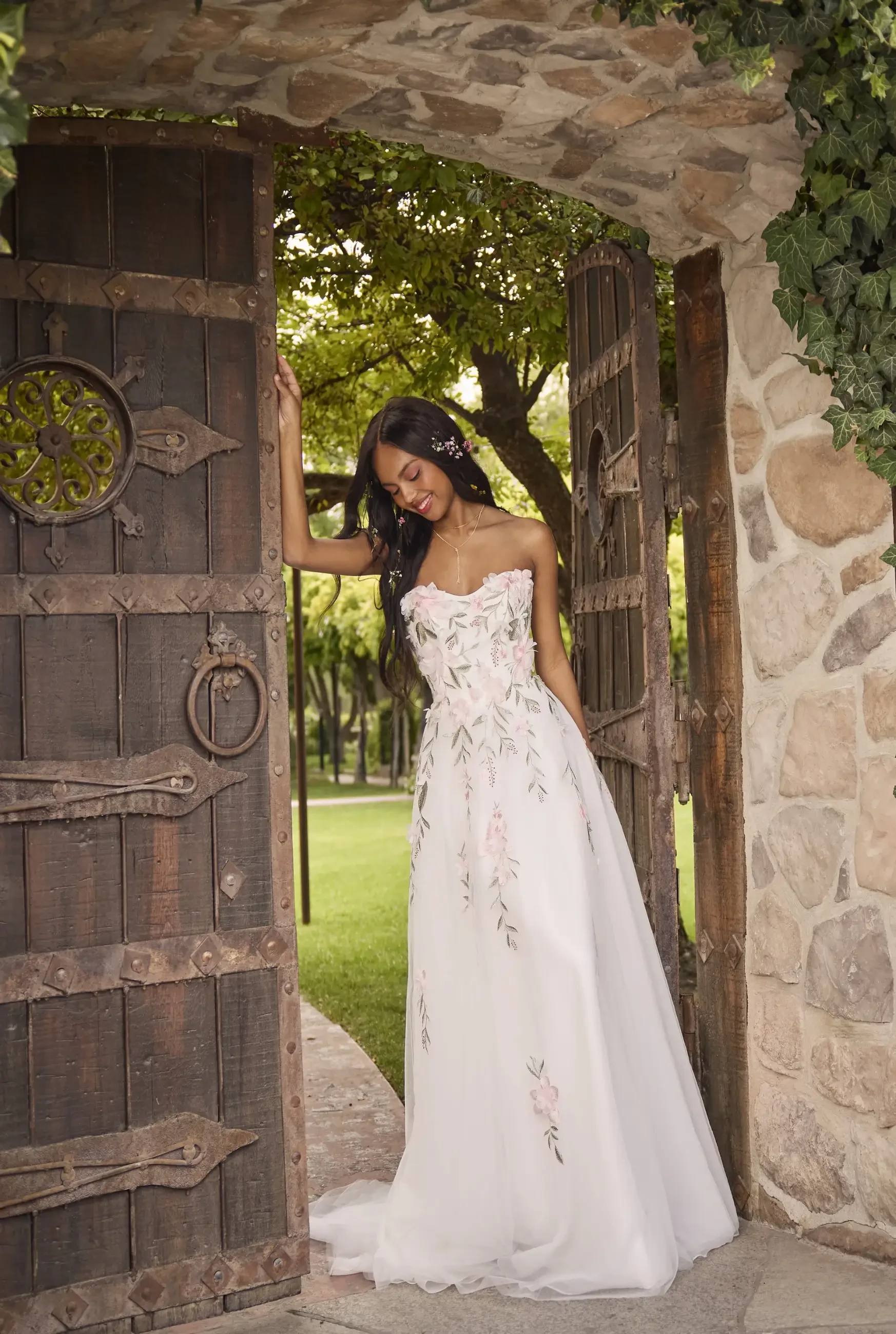 A woman in a strapless wedding dress with floral embroidery stands by an open stone gate, surrounded by greenery.