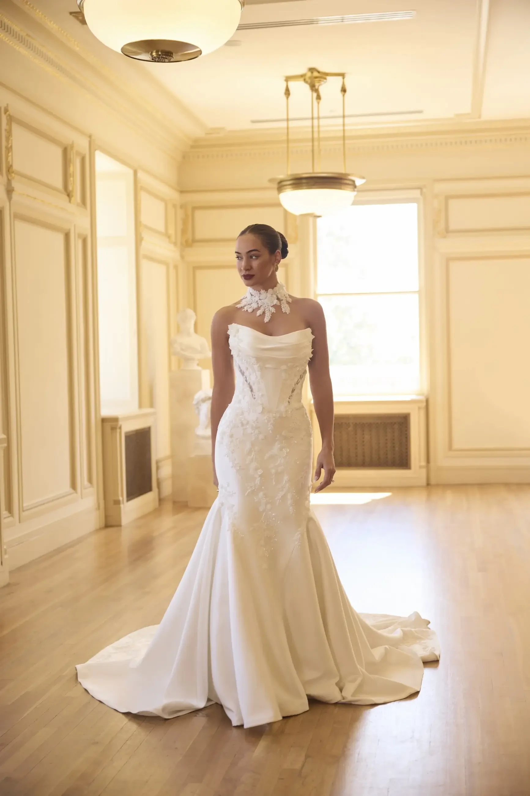 A woman in a white wedding dress stands elegantly in a well-lit, ornate room with wooden flooring and decorative moldings.