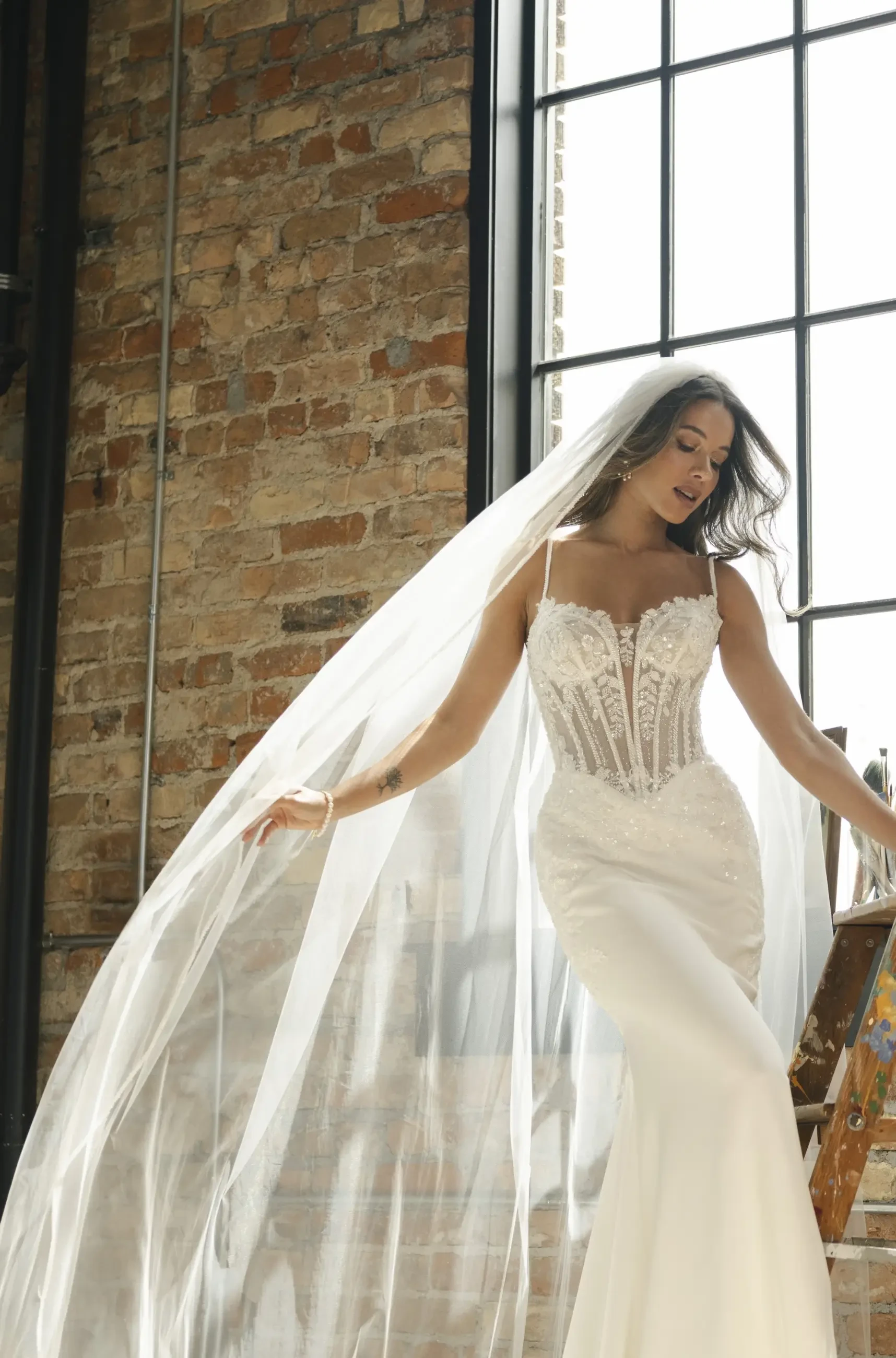 A woman in a lace corset bridal gown stands by a sunlit window. Her long veil flows elegantly, contrasting with the exposed brick wall background.