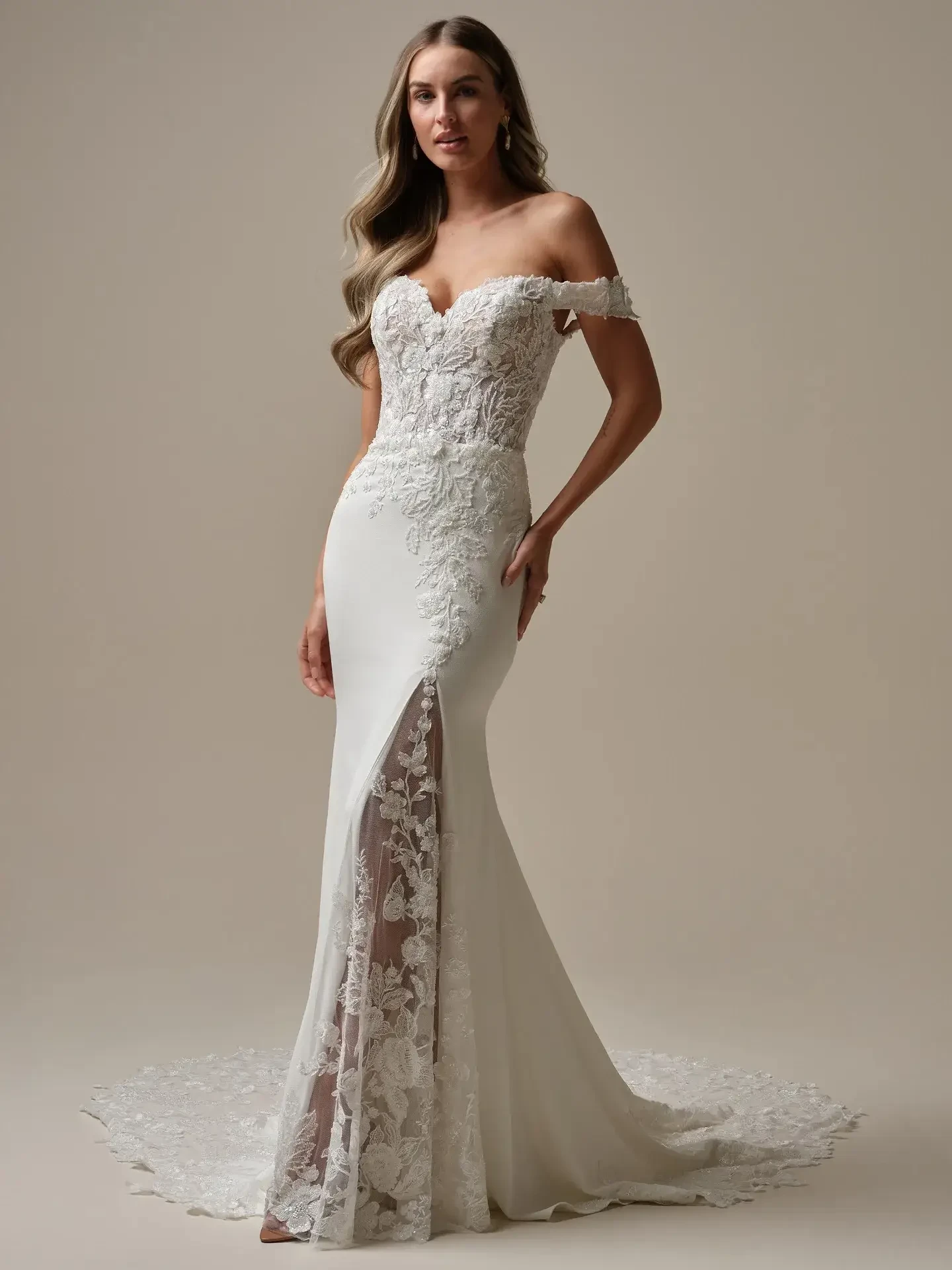 Model is wearing an off the shoulder lace fit and flare bridal gown with a lace inset on the high slit