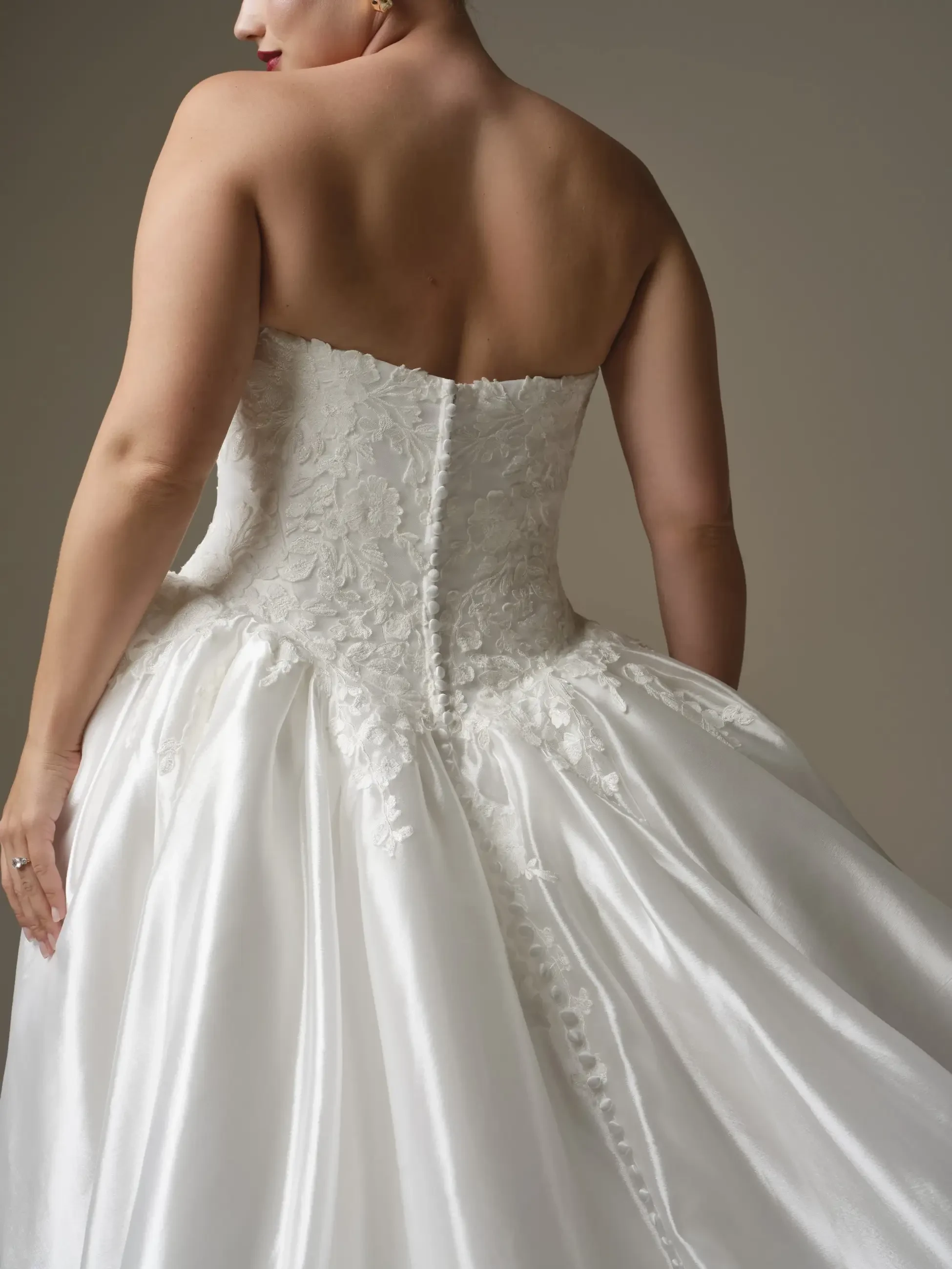 A woman is seen from behind, wearing a strapless white wedding dress with intricate lace detailing and a flowing skirt.