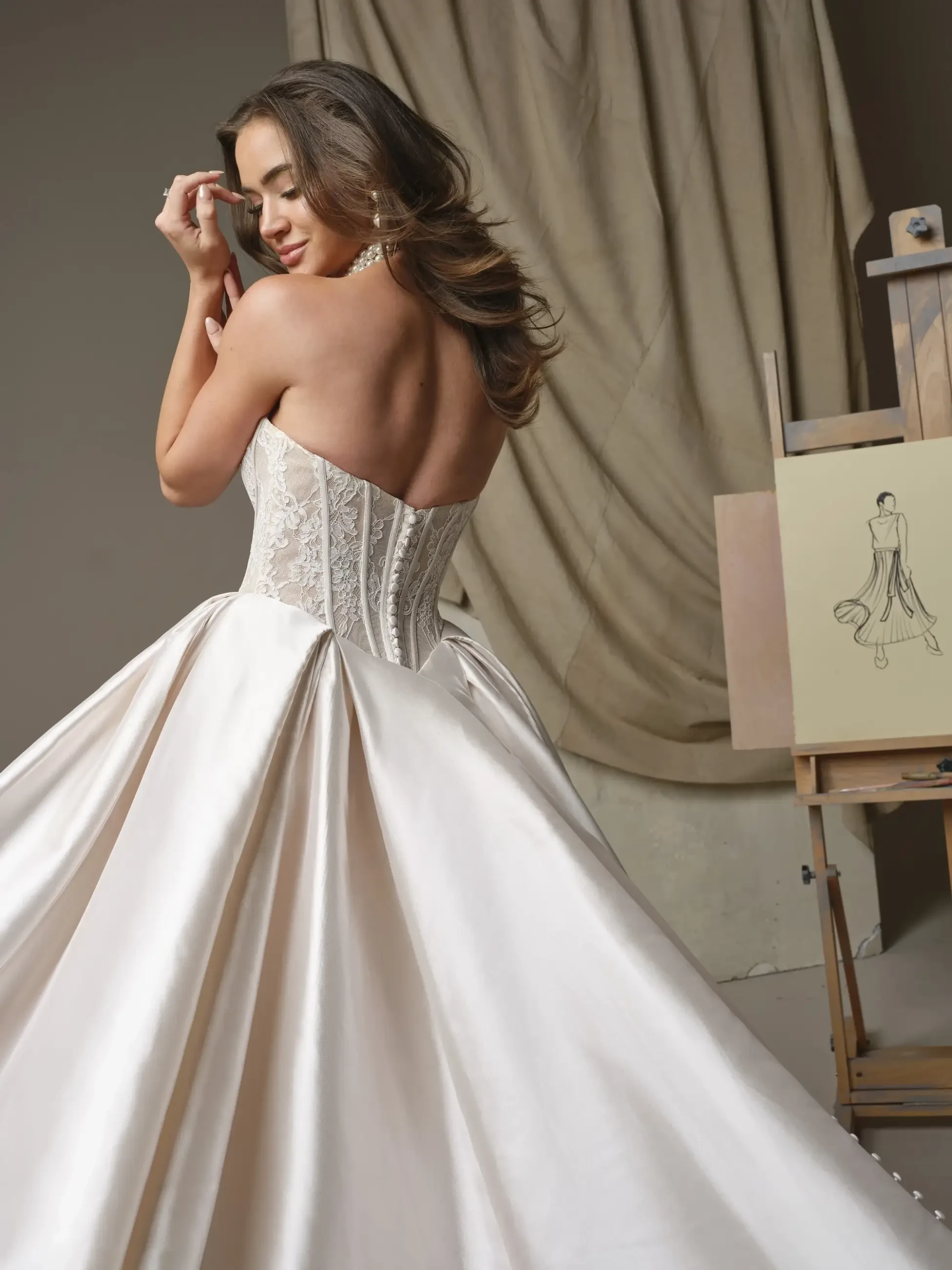 A person in a strapless, lace corset ball gown poses gracefully, eyes closed in delight. An elegant dress sketch rests on an easel nearby, evoking a serene, artistic vibe.