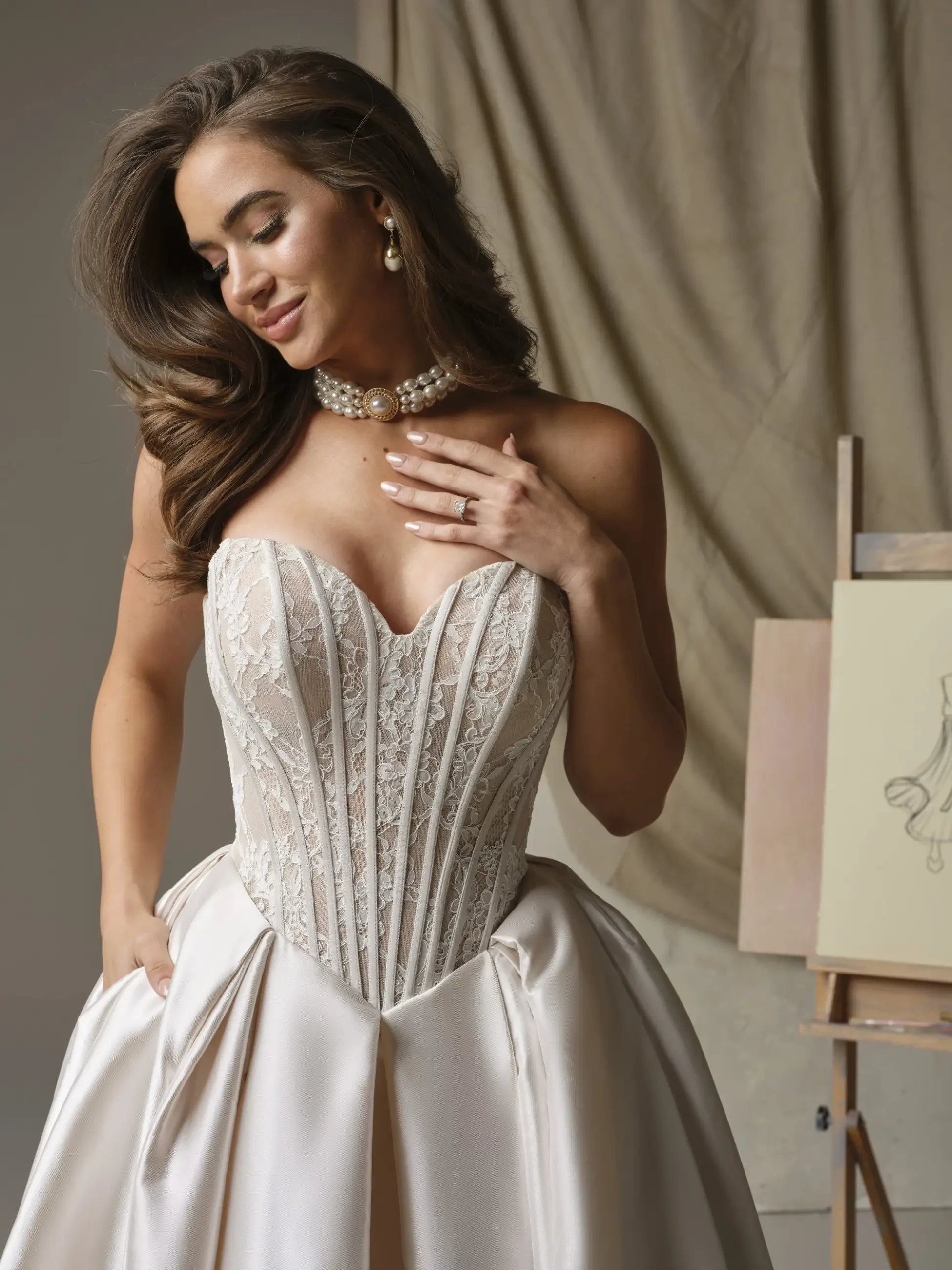 Woman in an elegant gown with lace corset, looking down, touching her chest. Wearing pearl jewelry. Art sketch on an easel in the background.
