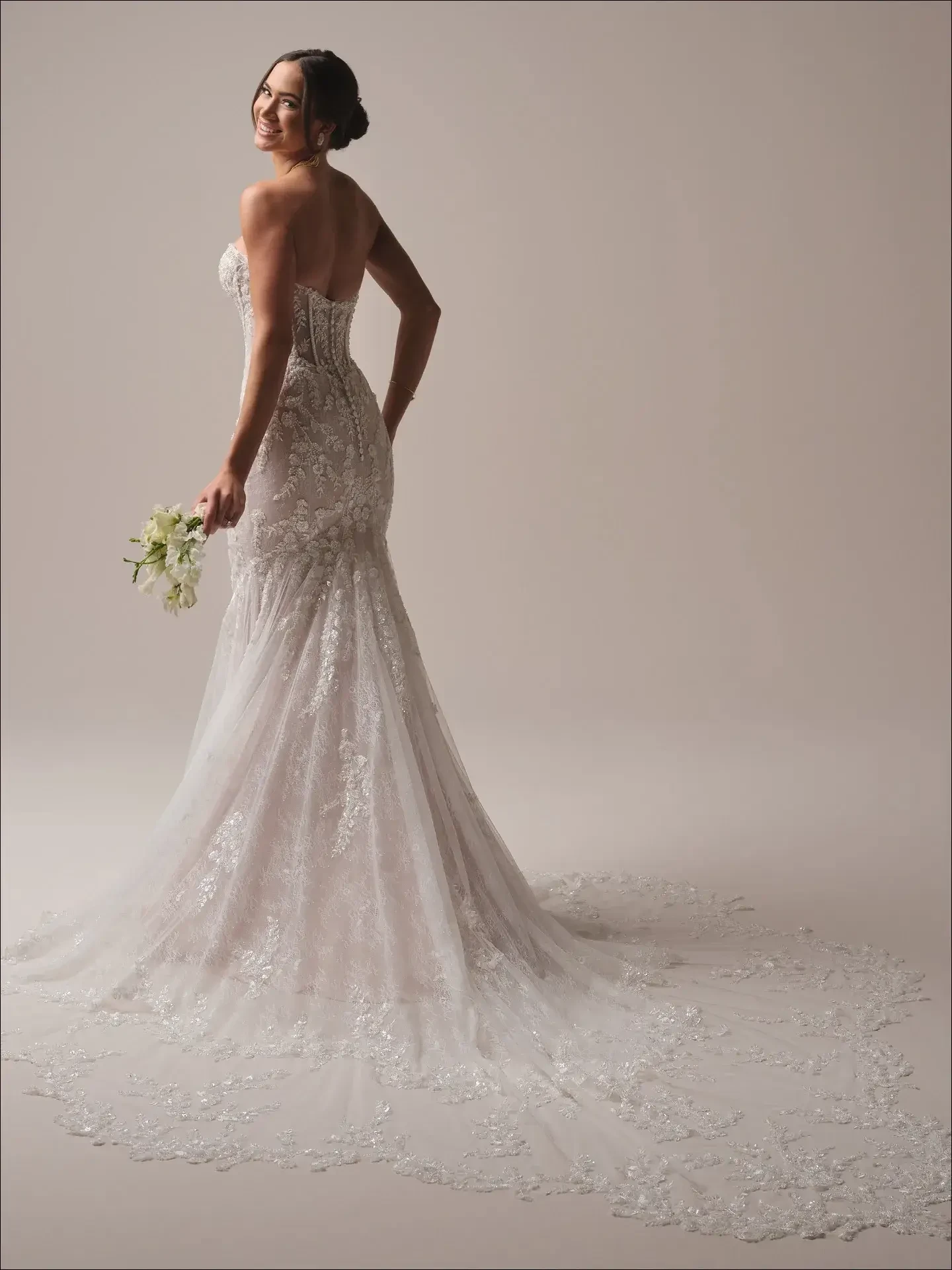Model is wearing a mermaid gown with a plunging illusion sweetheart neckline, glitter illusion bodice, and scalloped lace train.