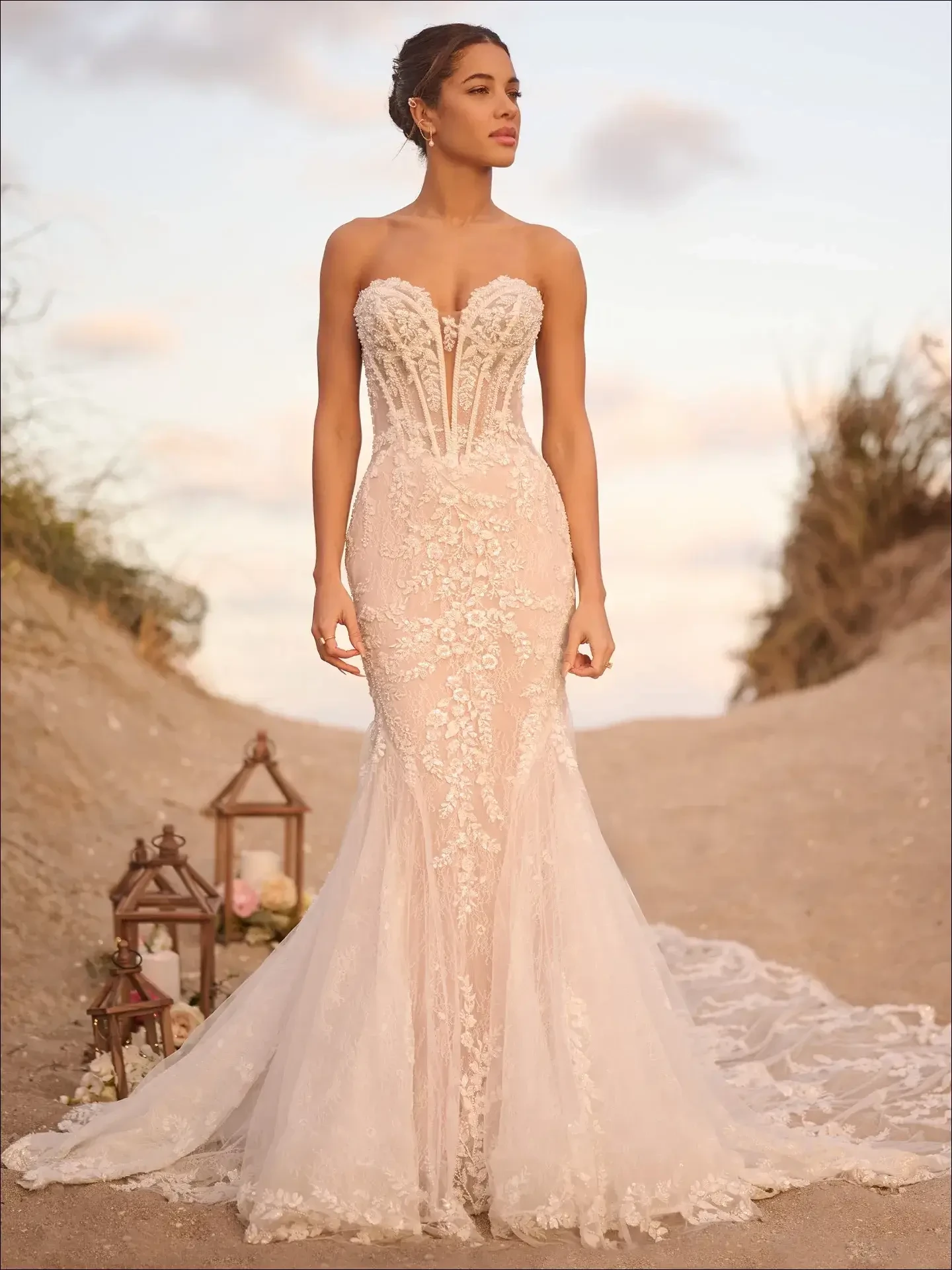 Model is wearing a mermaid gown with a plunging illusion sweetheart neckline, glitter illusion bodice, and scalloped lace train.