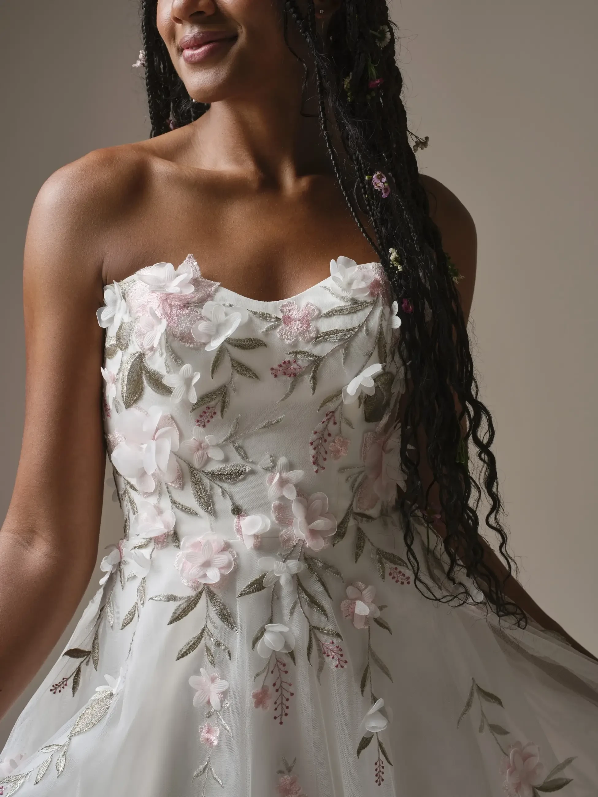 A close-up of a woman wearing a strapless, floral-embroidered dress with intricate designs in soft pastel colors. Her long, braided hair has small flowers intertwined, and she is smiling gently.
