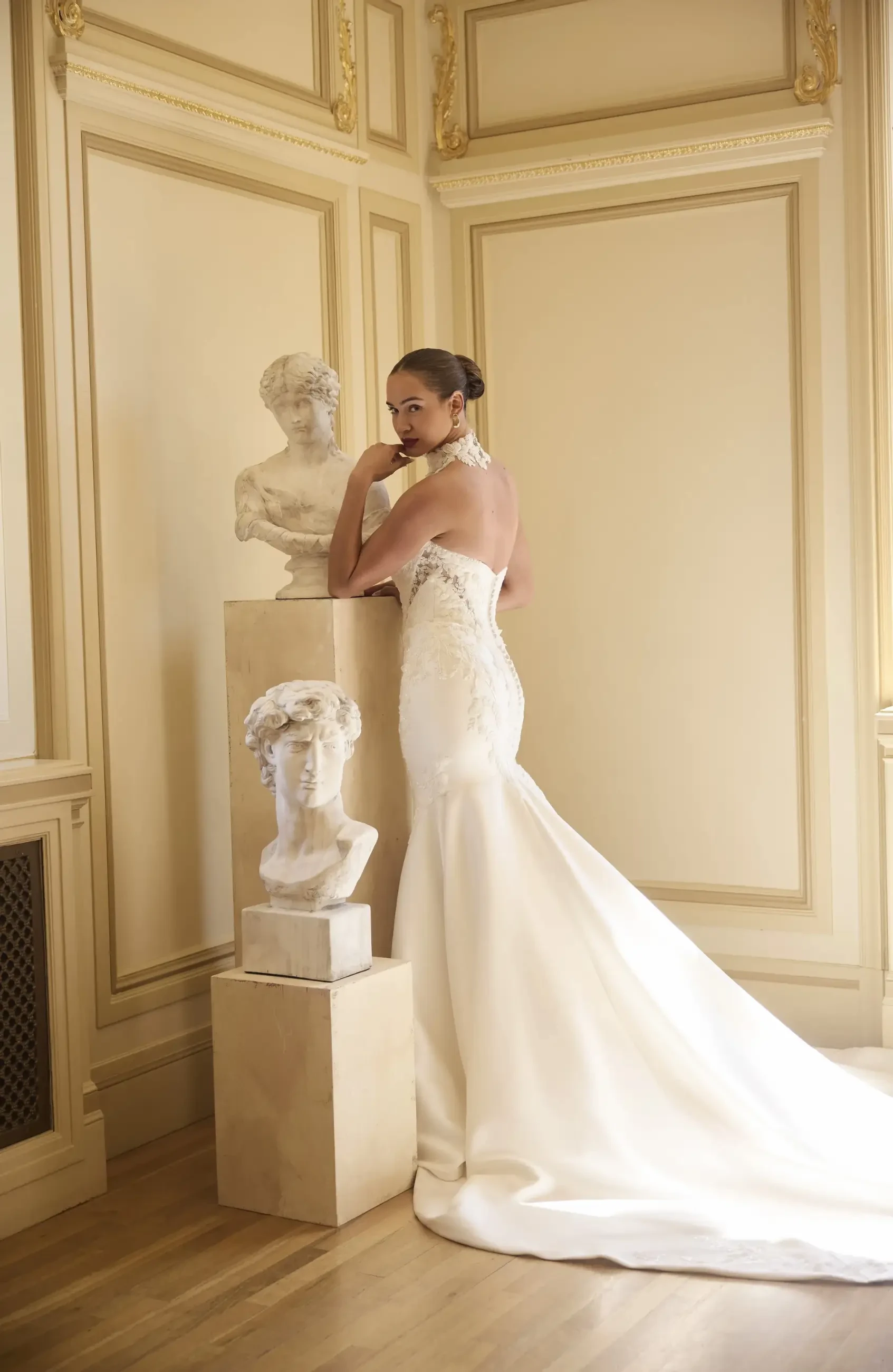A woman in an elegant strapless white gown stands beside classical bust sculptures, within an ornate room with paneled walls and golden accents.