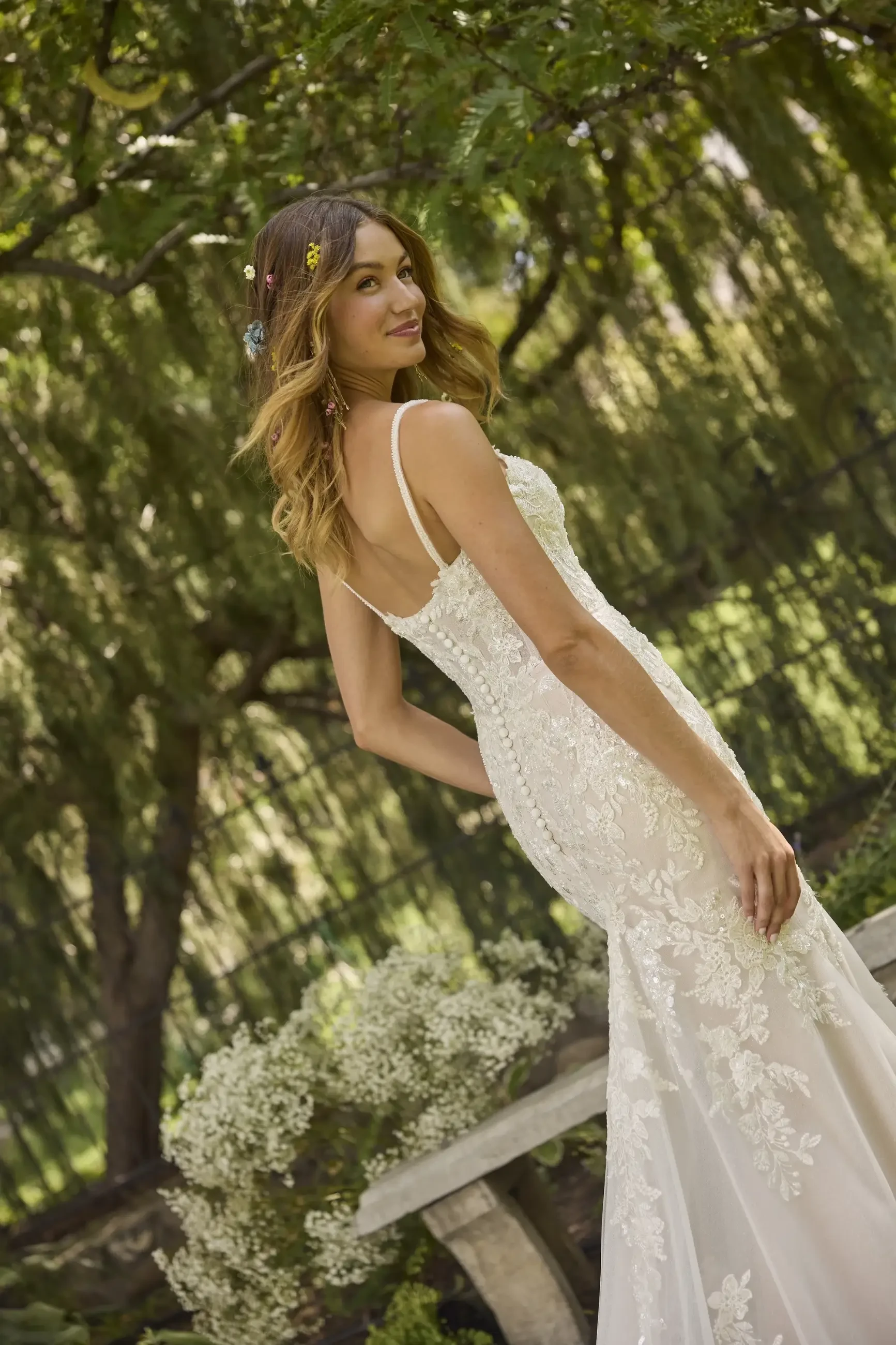 A woman in a lace wedding dress smiles joyfully amidst a lush garden setting. Sunlight filters through the trees, creating a serene, romantic atmosphere.