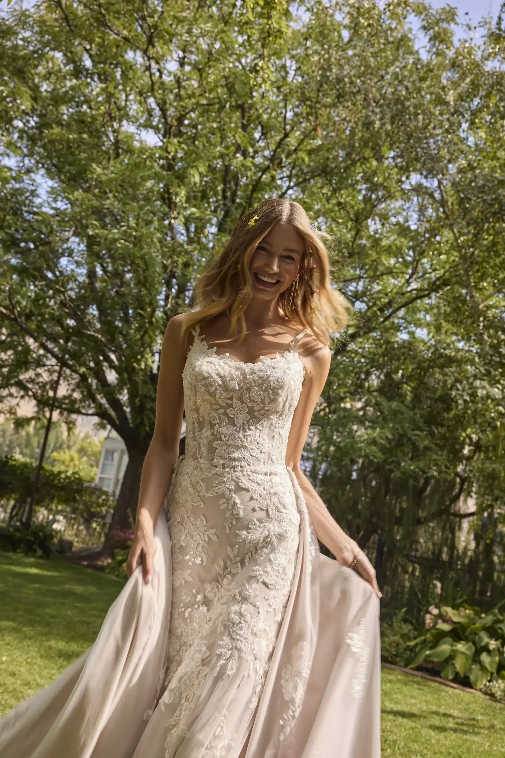 A woman in a lace wedding dress smiles joyfully amidst a lush garden setting. Sunlight filters through the trees, creating a serene, romantic atmosphere.