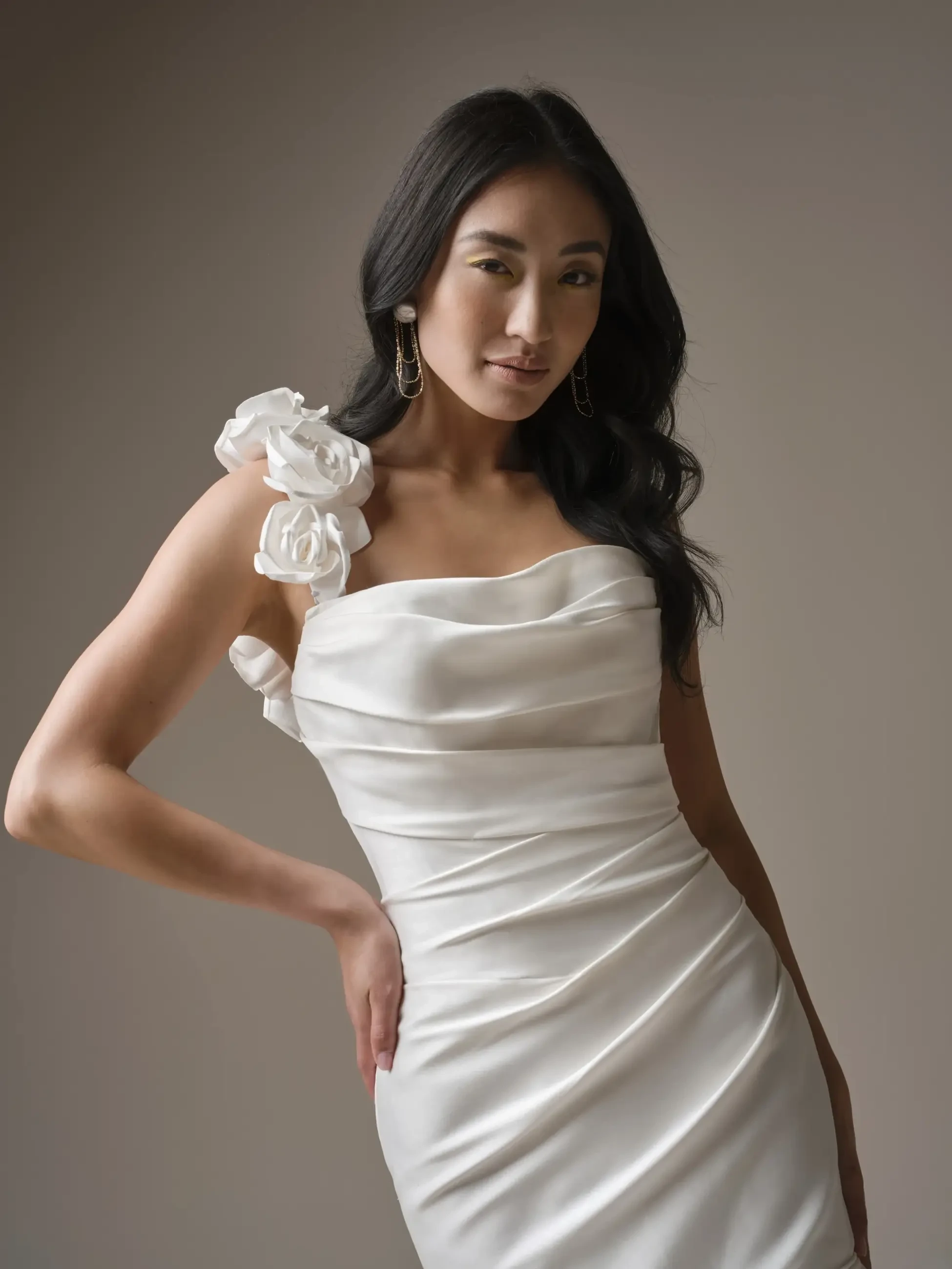 A person wearing a white dress with floral embellishments on one shoulder, posing confidently against a neutral background.