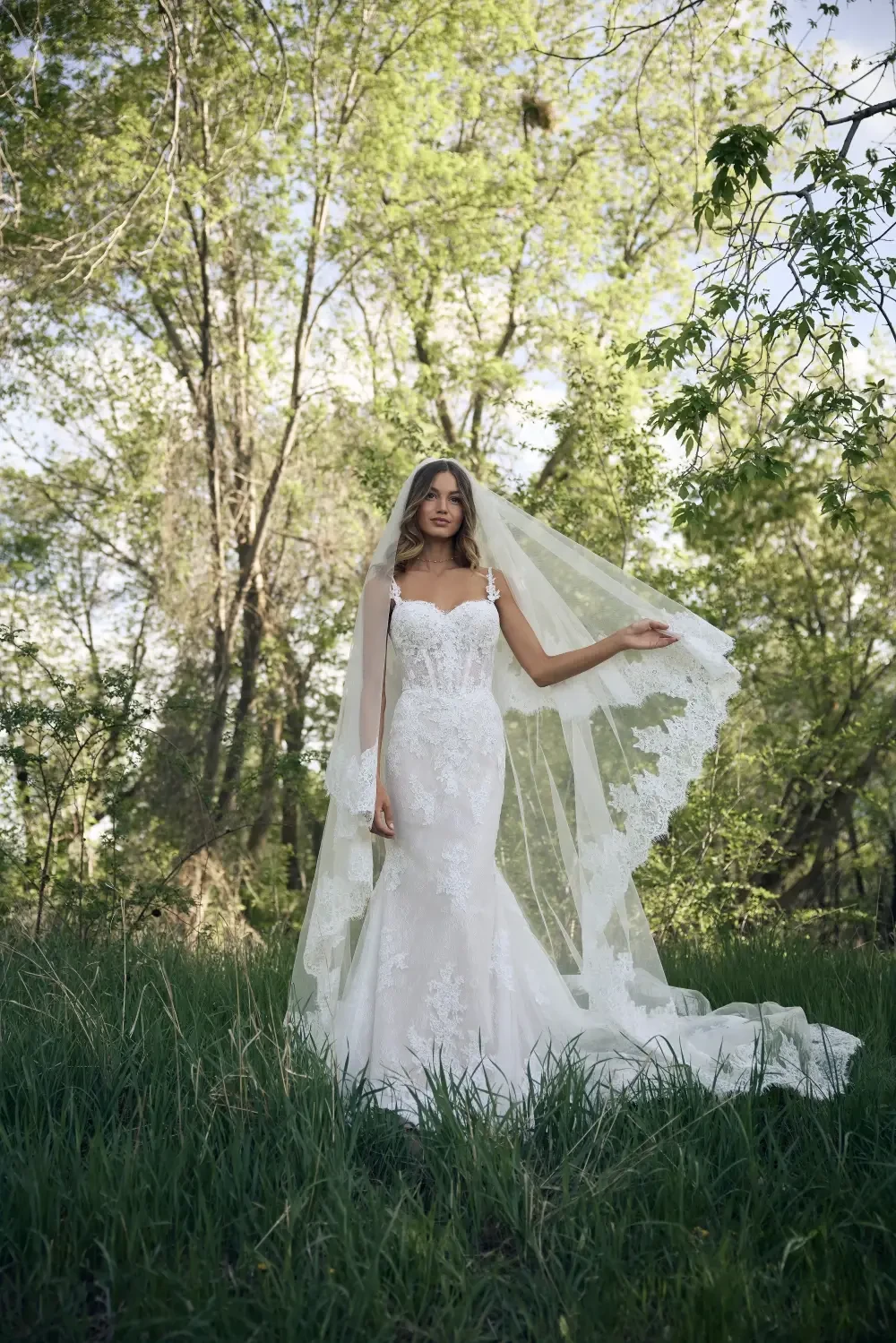 A bride in a white lace wedding dress stands in a green forest setting, gracefully holding a veil.