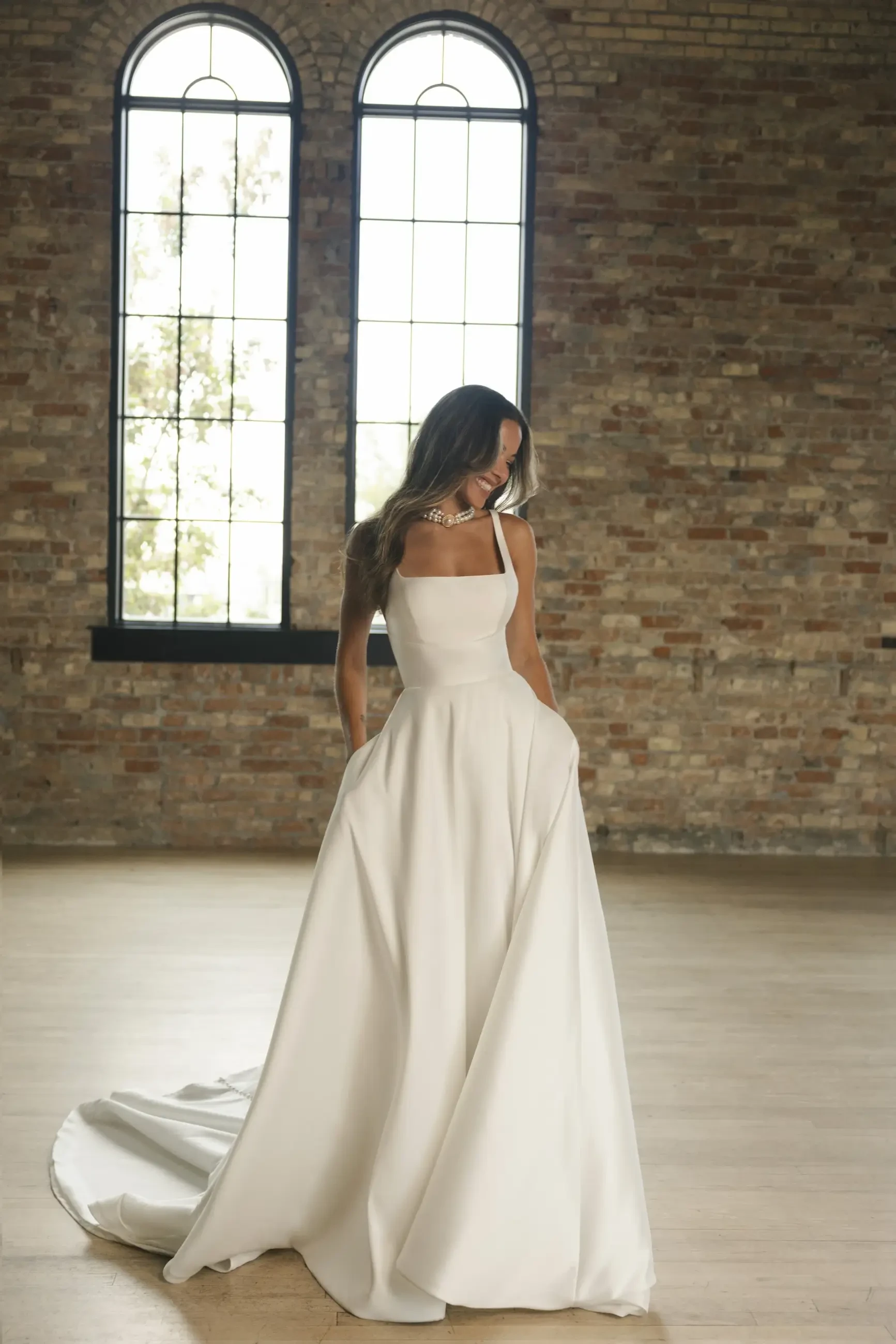 A person in a elegant white gown standing indoors with large windows and brick walls.