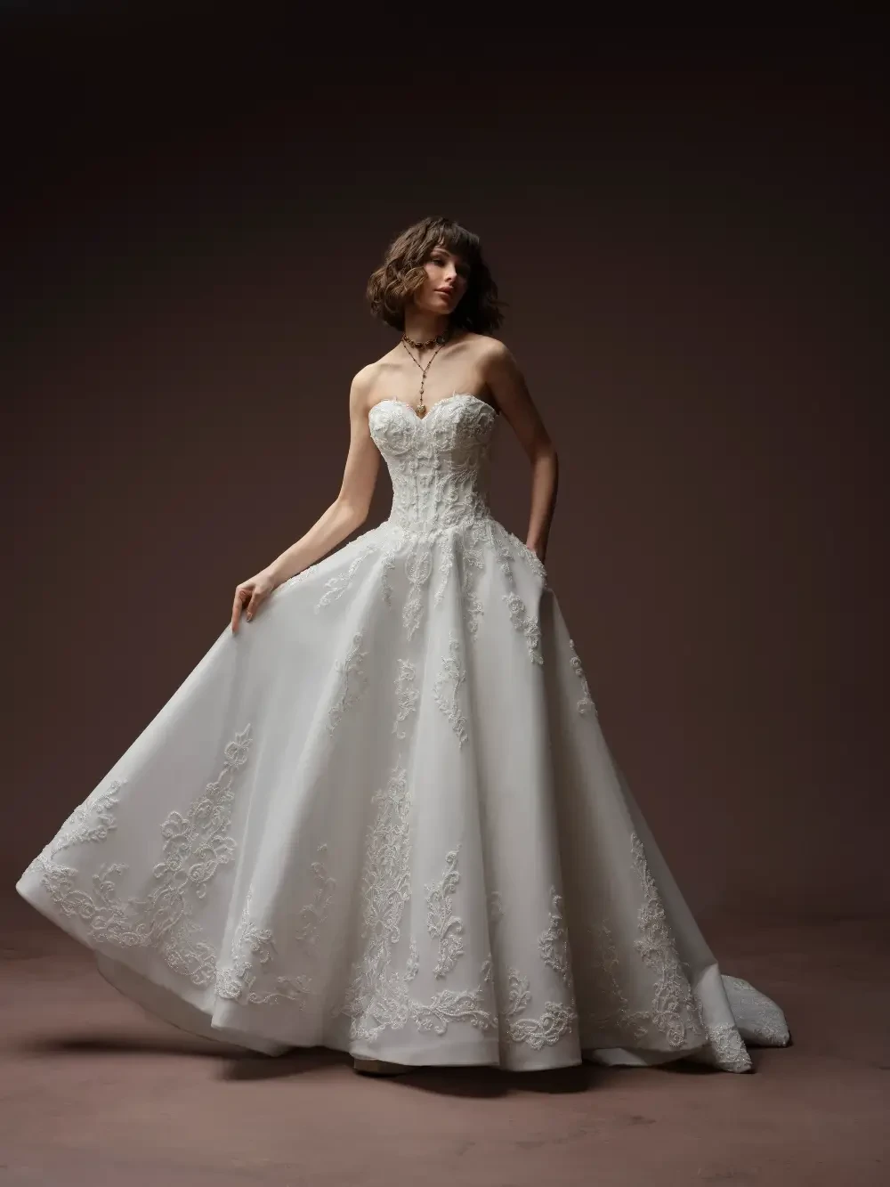 A model wearing a strapless white wedding gown with intricate lace detailing, standing gracefully against a dark background.