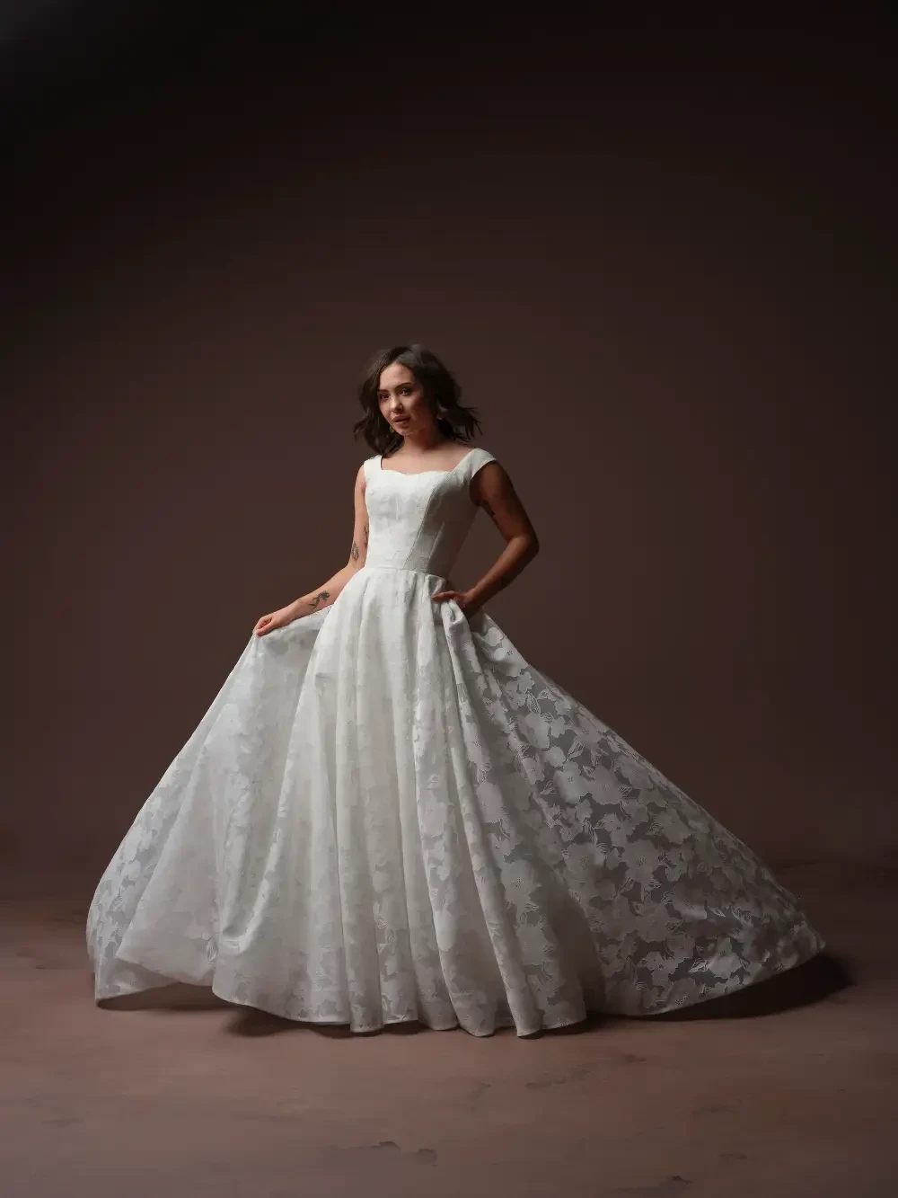 A model wearing a long, elegant white gown with a lace overlay, posed against a dark background.