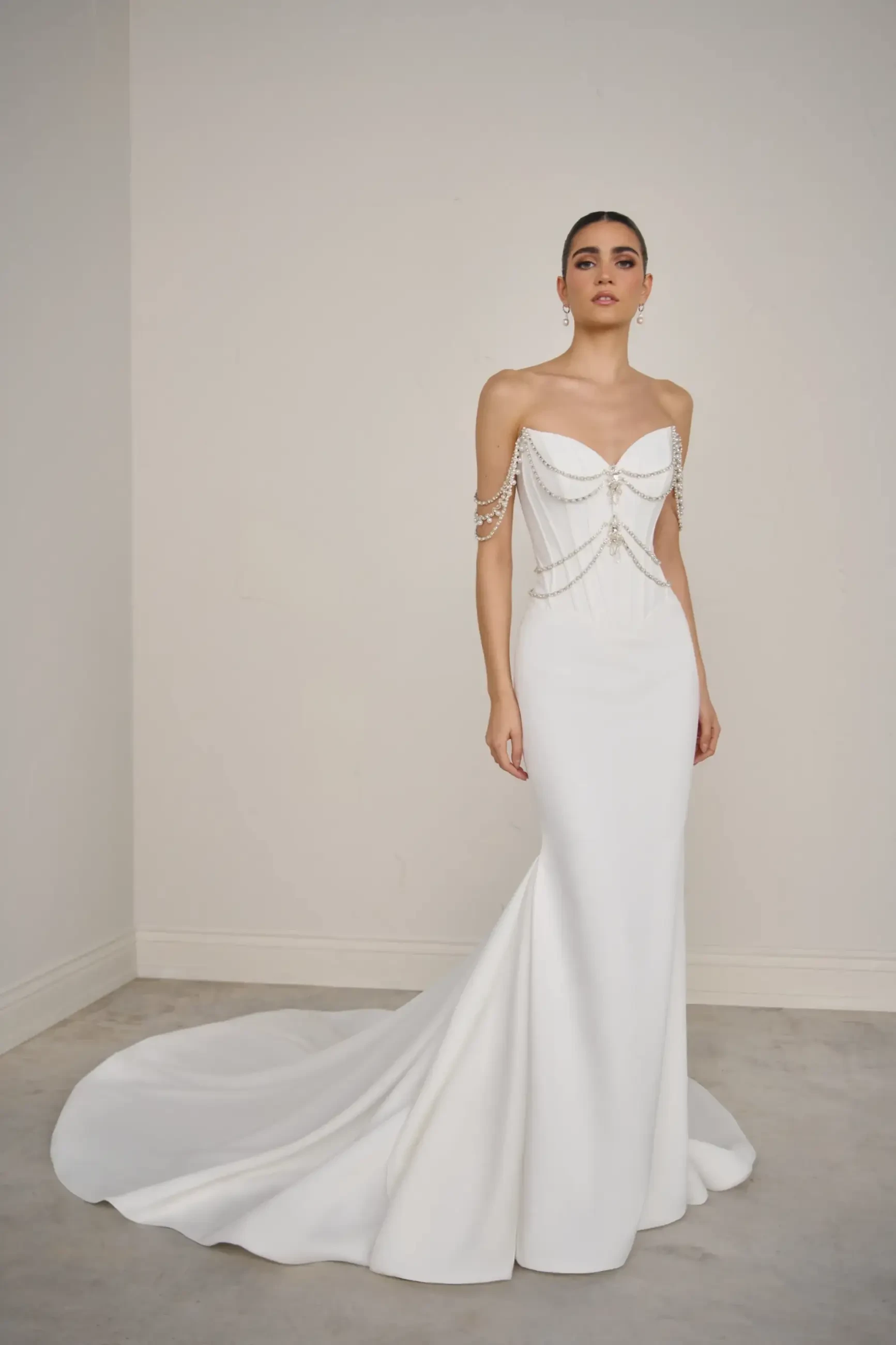 A woman stands confidently in a sleek white wedding dress with a long train. The gown features off-the-shoulder beaded straps, exuding elegance.