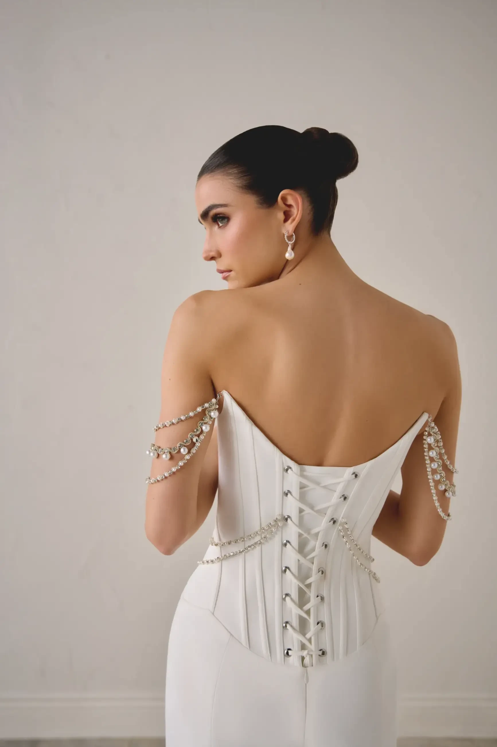 A woman in an elegant white dress with a lace-up back stands against a neutral background. Her hair is styled in a sleek bun, and she wears delicate pearl earrings and jeweled arm chains, conveying a sense of sophistication and grace.