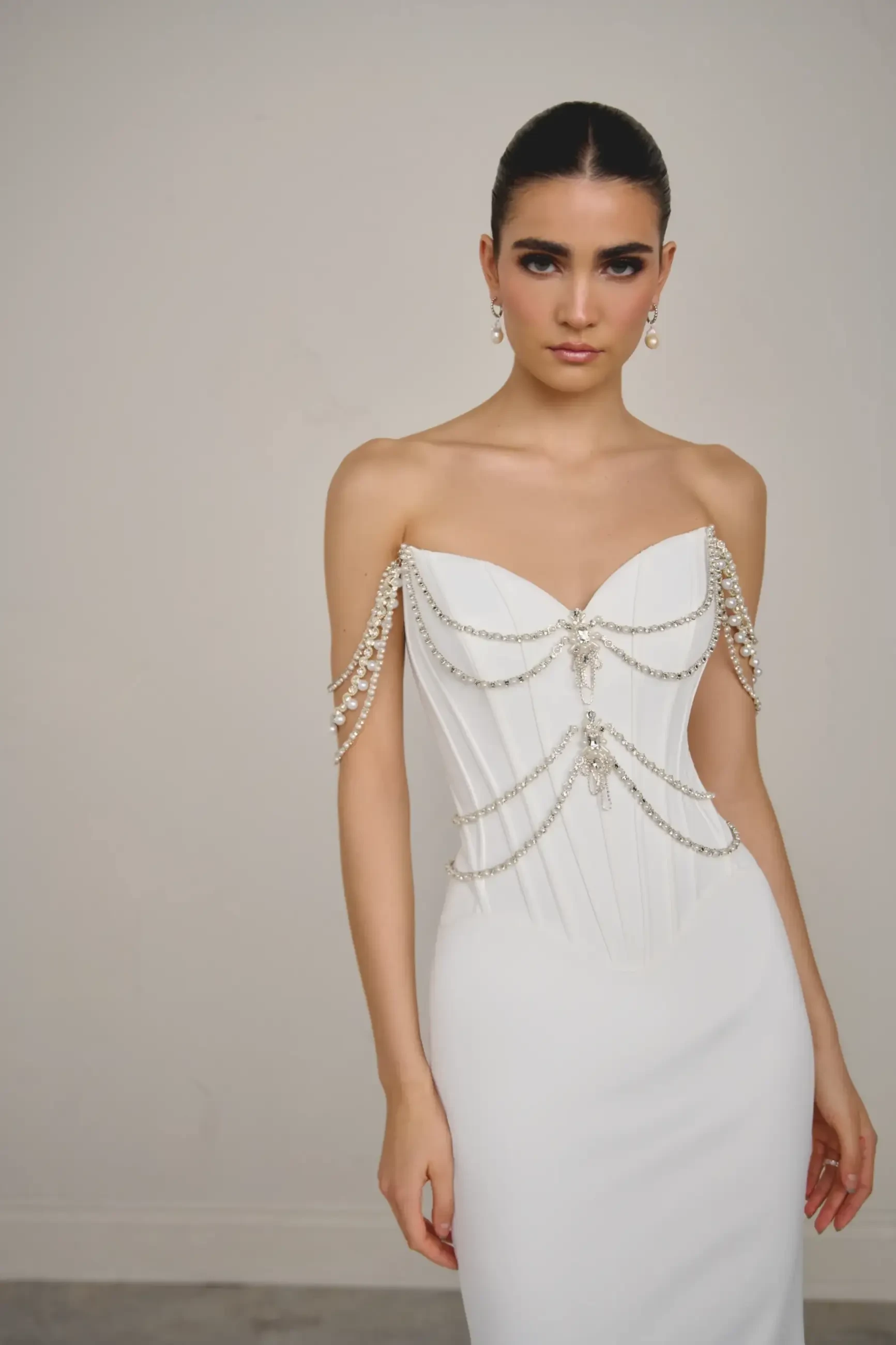 A model wearing a white strapless dress with beaded chains and embellishments, posing against a neutral background.
