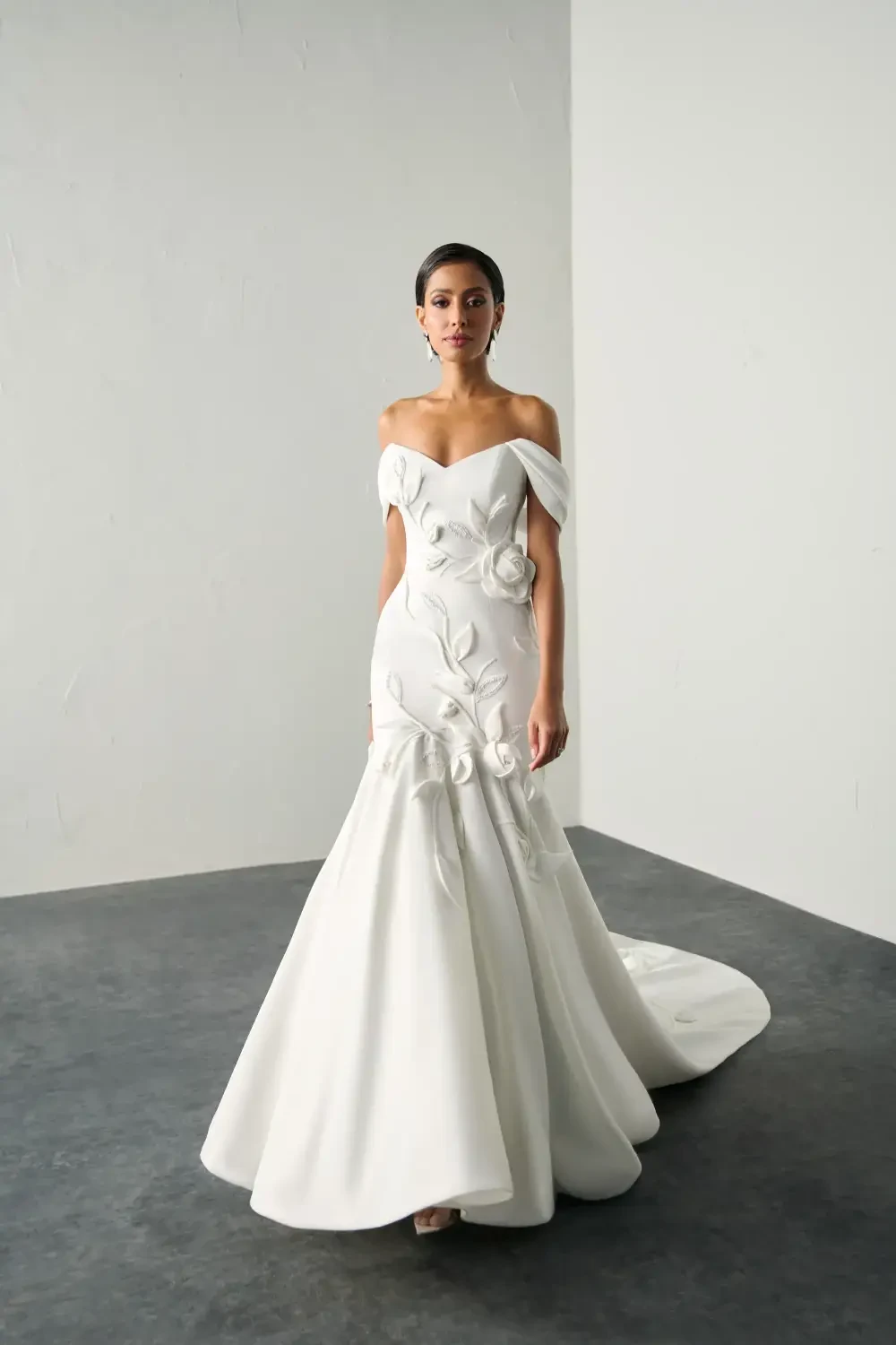A model wearing an elegant off-the-shoulder white wedding dress adorned with floral details, standing against a minimalistic background.
